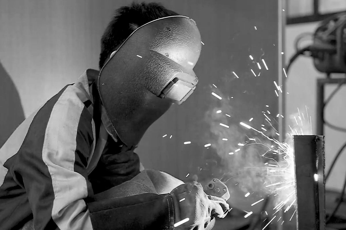 Worker using an angle grinder to cut metal, creating sparks while wearing a protective welding helmet.