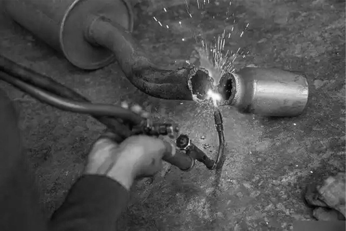 Worker using traditional torch welding to cut or repair a metal exhaust pipe with visible sparks.